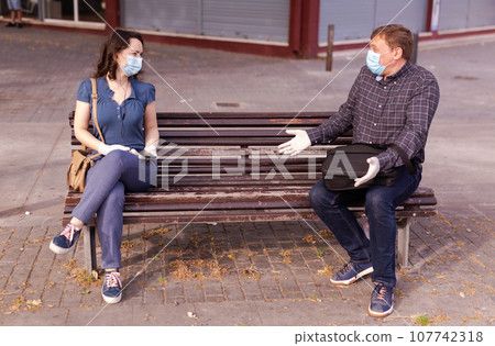 Emotional couple in protective medical masks against coronavirus communicate sittig on bench 107742318