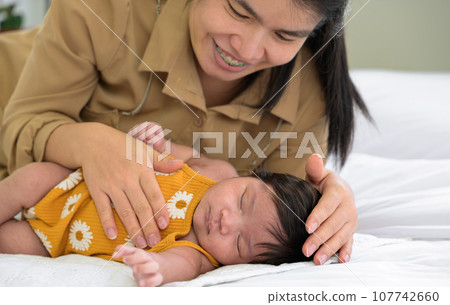 Smiling mother and child on white bed. Asian mom nursing her newborn baby girl at home. Smiling mother and child on white bed. Asian mom nursing her newborn baby girl at home. 107742660