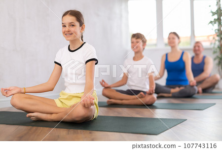 Pleasant preteen girl practicing lotus pose of yoga with her family Pleasant preteen girl practicing lotus pose of yoga with her family 107743116