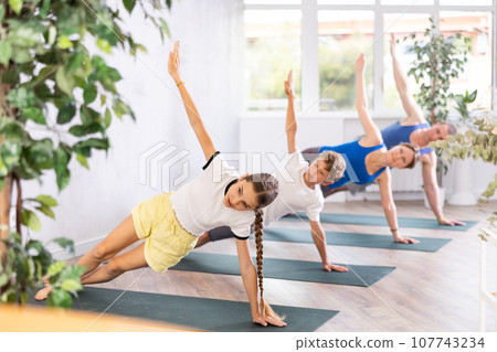 Pleasant preteen girl practicing side plank pose of yoga with her family 107743234