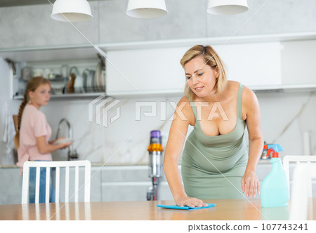 Blurry teen girl help mother doing housework in kitchen, mom cleaning dining table Blurry teen girl help mother doing housework in kitchen, mom cleaning dining table 107743241