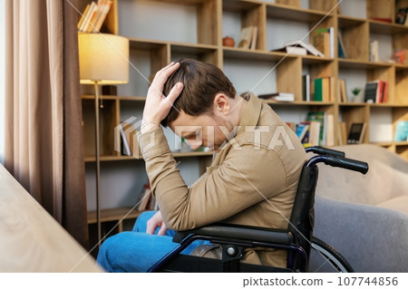 A young handsome man in a wheelchair looks out the window with a sad look A young handsome man in a wheelchair looks out the window with a sad look 107744856