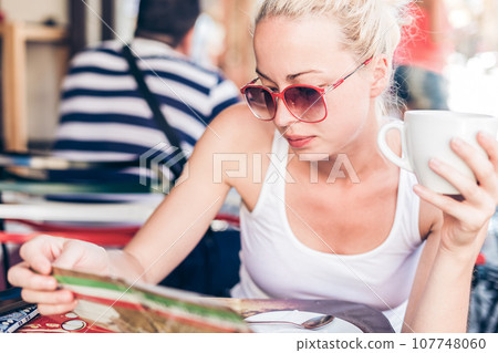 Calm casual blond lady wearing fashionable sunglasses, enjoying cup of coffee while checking menu in typical italian street coffee house or restaurant on warm summer day. Calm casual blond lady wearing fashionable sunglasses, enjoying cup of coffee while checking menu in typical italian street coffee house or restaurant on warm summer day. 107748060