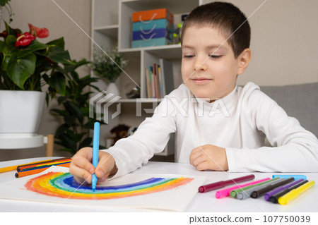 A small child draws a rainbow with felt-tip pens in a sketchbook while sitting at a table 107750039