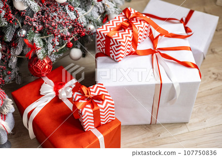 A variety of colored Christmas gifts stand near a decorative Christmas tree in the house on the floor under the branches. A variety of colored Christmas gifts stand near a decorative Christmas tree in the house on the floor under the branches. 107750836
