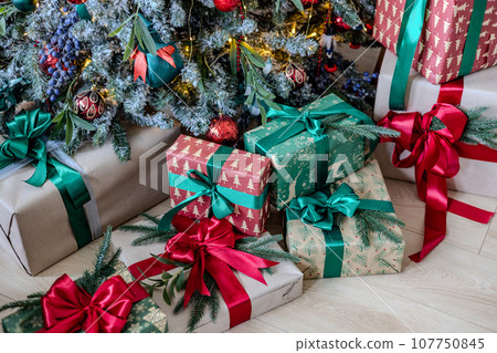 A variety of colored Christmas gifts stand near a decorative Christmas tree in the house on the floor under the branches. 107750845