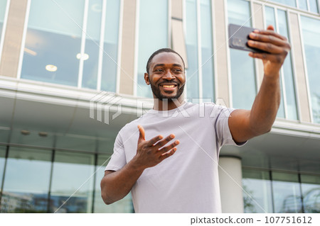 African American man holding smartphone having video chat on urban street in city. Guy blogger chatting with best friends in social network. Man having virtual meeting online chat video call outdoor African American man holding smartphone having video chat on urban street in city. Guy blogger chatting with best friends in social network. Man having virtual meeting online chat video call outdoor 107751612