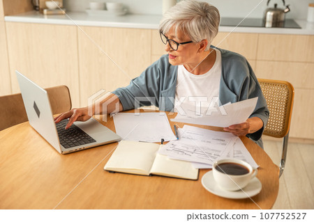 Middle aged senior woman sit with laptop and paper document, Pensive older mature lady reading paper bill pay online at home managing bank finances calculating taxes planning loan debt pension payment Middle aged senior woman sit with laptop and paper document, Pensive older mature lady reading paper bill pay online at home managing bank finances calculating taxes planning loan debt pension payment 107752527