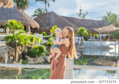 Amidst tropical palms and thatched roofs, a loving mom embraces her baby, sharing warmth and affection in a tranquil outdoor setting 107754374