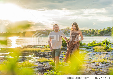 A happy, mature couple over 40, enjoying a leisurely walk on the waterfront On the Sunset, their joy evident as they embrace the journey of pregnancy later in life 107754503