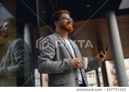 Handsome male freelancer holding phone while standing near office during break time and drink coffee 107755102