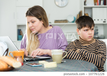 Young mother working at laptop and unhappy preteen son Young mother working at laptop and unhappy preteen son 107761226