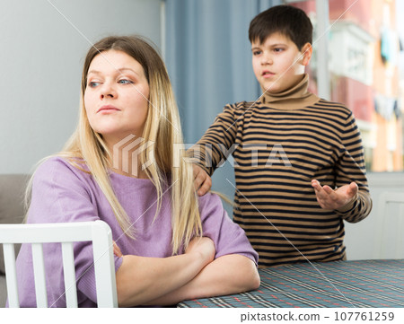 Upset mother sitting at table after arguing with son indoors 107761259
