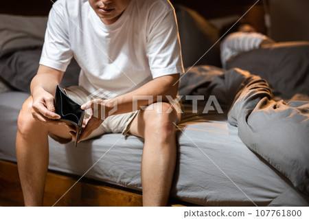 Stressed asian man holding empty wallet,sitting on bed at home,debt burden,housing loan repayment problems,financial hardship,be short of money,house installment liabilities,debt restructuring concept 107761800