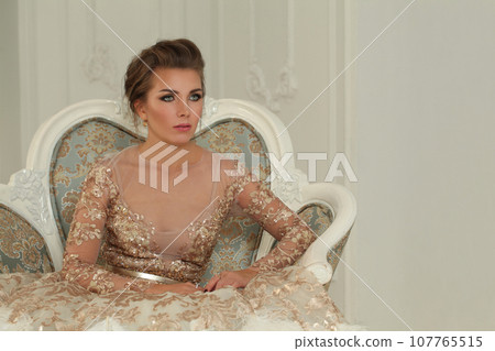 Perfect stylish woman wearing gold evening gown sitting on armchair in luxurious interior 107765515