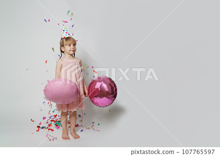 Happy little child girl in birthday hat having fun, holding balloon and standing in confetti rain Happy little child girl in birthday hat having fun, holding balloon and standing in confetti rain 107765597
