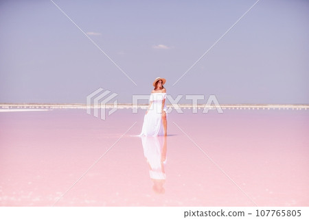 Woman in pink salt lake. She in a white dress and hat enjoys the scenic view of a pink salt lake as she walks along the white, salty shore, creating a lasting memory. Woman in pink salt lake. She in a white dress and hat enjoys the scenic view of a pink salt lake as she walks along the white, salty shore, creating a lasting memory. 107765805