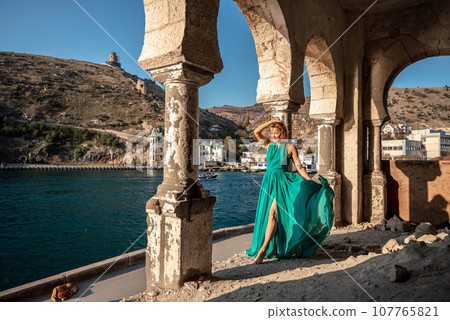 Woman dress sea columns. Rear view of a happy blonde woman in a long mint dress posing against the backdrop of the sea in an old building with columns. 107765821