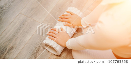 Adult athletic woman, in white bodysuit, performing fascia exercises on the floor - caucasian woman using a massage foam roller - a tool to relieve tension in the back and relieve muscle pain - the Adult athletic woman, in white bodysuit, performing fascia exercises on the floor - caucasian woman using a massage foam roller - a tool to relieve tension in the back and relieve muscle pain - the 107765868