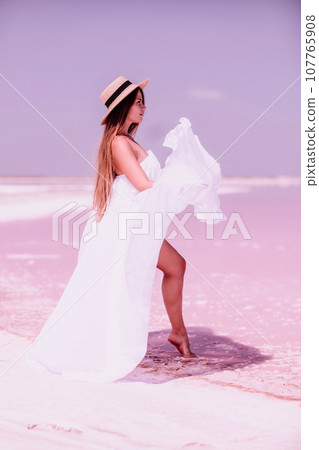 Woman in pink salt lake. She in a white dress and hat enjoys the scenic view of a pink salt lake as she walks along the white, salty shore, creating a lasting memory. 107765908
