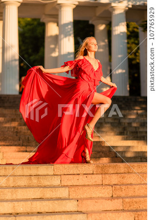 A woman in a long red dress against the backdrop of sunrise, bright golden light of the sun's rays. The concept of femininity, harmony. 107765929