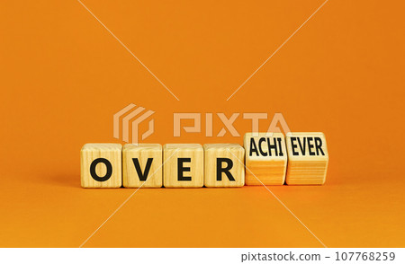 Achiever or overachiever symbol. Businessman turns wooden cubes and changes word Achiever to Overachiever. Beautiful orange background. Business achiever or overachiever concept. Copy space. 107768259