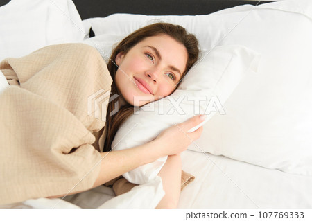 Gorgeous woman resting in bed wearing pyjamas, hugging her pillow and smiling 107769333