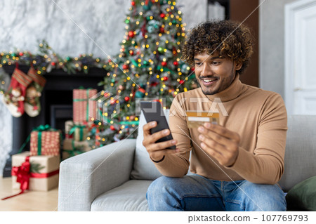 Young man on Christmas at home in the living room sitting near the Christmas tree decorated with Christmas decorations, hispanic smiling holding a bank credit card and phone, choosing gifts online. 107769793