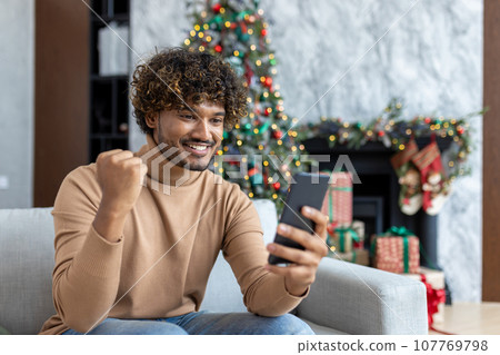 Successful man received online notification of victory win, winner for christmas sitting near tree living room on sofa in house indoors, hispanic using app on smartphone, holding hand up achievement. Successful man received online notification of victory win, winner for christmas sitting near tree living room on sofa in house indoors, hispanic using app on smartphone, holding hand up achievement. 107769798