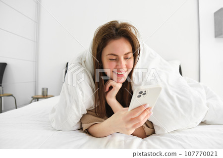 Portrait of gorgeous girl wakes up in morning, lying in bed with smartphone, covered in blankets, reading social media feed on telephone, resting in bedroom Portrait of gorgeous girl wakes up in morning, lying in bed with smartphone, covered in blankets, reading social media feed on telephone, resting in bedroom 107770021