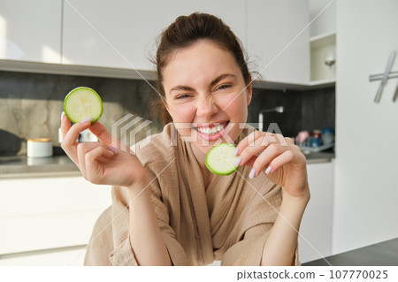Portrait of beautiful brunette girl cooking in the kitchen, posing in bathrobe at home, holding zucchini, showing happy smile, making healthy food, vegetarian meal Portrait of beautiful brunette girl cooking in the kitchen, posing in bathrobe at home, holding zucchini, showing happy smile, making healthy food, vegetarian meal 107770025