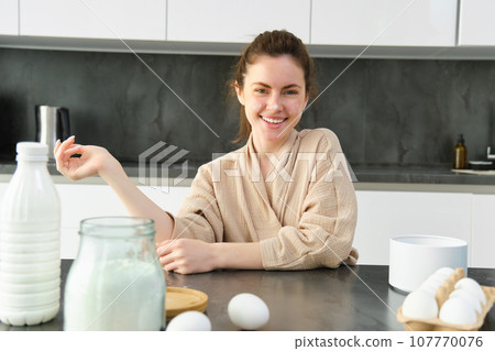 Attractive young cheerful girl baking at the kitchen, making dough, holding recipe book, having ideas 107770076
