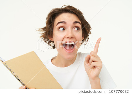 Portrait of happy young woman with planner, student with homework notes, pointing at upper right corner, showing banner and making amazed face expression, standing over white background Portrait of happy young woman with planner, student with homework notes, pointing at upper right corner, showing banner and making amazed face expression, standing over white background 107770082