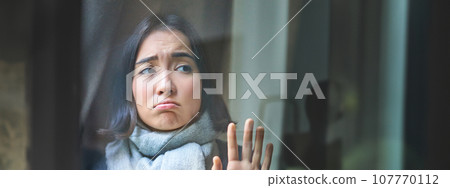 Vertical shot of upset asian woman looking outside, touching window, staring at street with gloomy jealous face, feeling ill, staying at home with covid or flu Vertical shot of upset asian woman looking outside, touching window, staring at street with gloomy jealous face, feeling ill, staying at home with covid or flu 107770112