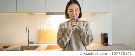 Portrait of smiling asian woman standing on her kitchen, drinking coffee and looking at camera, concept of cozy and comfortable home Portrait of smiling asian woman standing on her kitchen, drinking coffee and looking at camera, concept of cozy and comfortable home 107770141