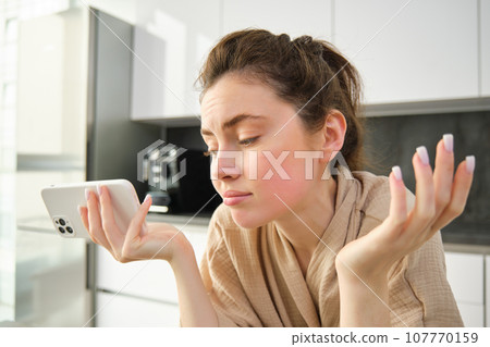 Attractive young cheerful girl baking at the kitchen, making dough, holding recipe book, having ideas 107770159