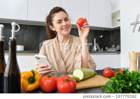 Young woman orders groceries on mobile app. Girl in bathrobe sits in the kitchen with vegetables, looking for recipe to cook dinner, using smartphone application 107770162