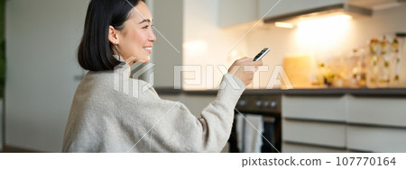 Close up portrait of smiling asian woman watching television, sitting on sofa with remote and switching chanel, looking relaxed 107770164