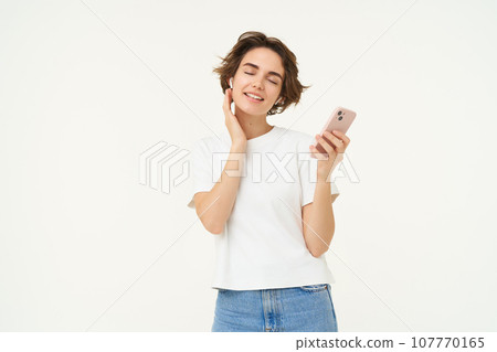 Portrait of smiling, beautiful young woman in wireless headphones, puts on music in earphones, holds smartphone, using streaming app, standing over white background 107770165