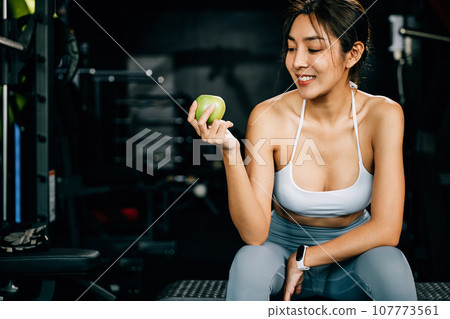An Asian girl holds a green apple in a fitness gym, emphasizing the importance of healthy food choices for maintaining a slim and sportive physique. Healthy fitness and eating lifestyle concept An Asian girl holds a green apple in a fitness gym, emphasizing the importance of healthy food choices for maintaining a slim and sportive physique. Healthy fitness and eating lifestyle concept 107773561