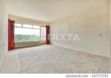 an empty living room with red drapes on the windows and carpeted floor in front of the window looking out 107773787