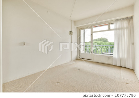 an empty living room with white walls and beige carpet, there is a window in the wall to the right an empty living room with white walls and beige carpet, there is a window in the wall to the right 107773796