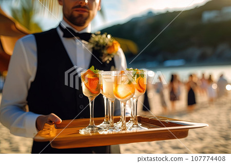 Waiter at a party, carrying a tray with champagne or wine glasses. Outdoor wedding, party on the beach Waiter at a party, carrying a tray with champagne or wine glasses. Outdoor wedding, party on the beach 107774408