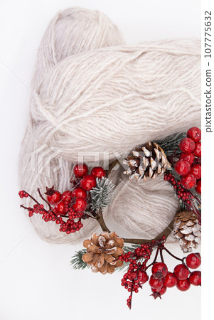 Gray woolen skeins or yarn of wool with branch of red berries and pine cones on white background. Handmade, knitting, hobby. Copy space 107775632