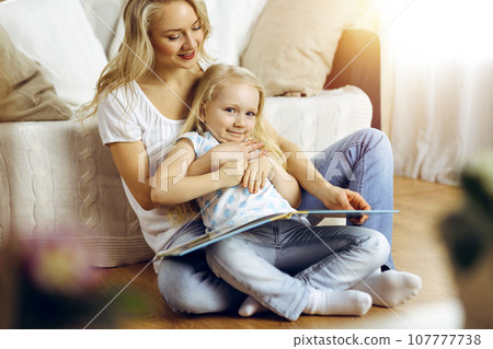 Happy family. Blonde young mother reading a book to her cute daughter while sitting at wooden floor in sunny room. Motherhood concept Happy family. Blonde young mother reading a book to her cute daughter while sitting at wooden floor in sunny room. Motherhood concept 107777738