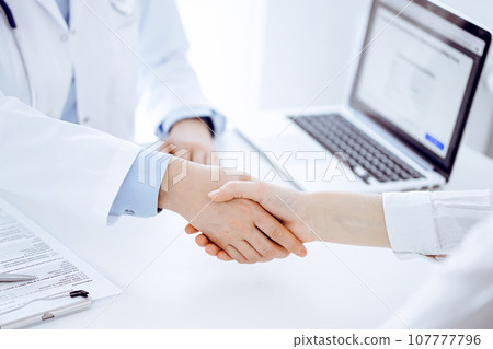 Doctor and patient shaking hands while sitting opposite of each other at the table in clinic, just hands close up. Medicine concept Doctor and patient shaking hands while sitting opposite of each other at the table in clinic, just hands close up. Medicine concept 107777796