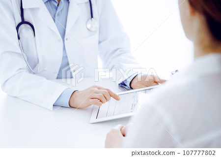Doctor and patient sitting opposite each other at the desk in clinic. The focus is on female physician's hands pointing into tablet computer touchpad, close up. Medicine concept 107777802