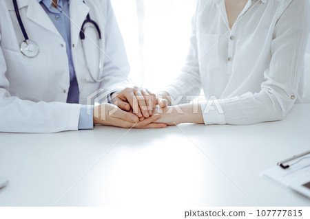 Doctor and patient sitting near each other at the table in clinic office. The focus is on female physician's hands reassuring woman, only hands, close up. Medicine concept Doctor and patient sitting near each other at the table in clinic office. The focus is on female physician's hands reassuring woman, only hands, close up. Medicine concept 107777815