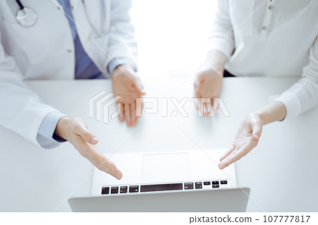 Doctor and patient sitting near each other at the table in clinic. The focus is on female hands pointing into tablet computer touchpad together. Medicine concept Doctor and patient sitting near each other at the table in clinic. The focus is on female hands pointing into tablet computer touchpad together. Medicine concept 107777817