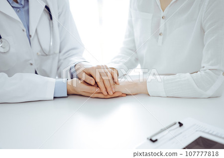 Doctor and patient sitting near each other at the table in clinic office. The focus is on female physician's hands reassuring woman, only hands, close up. Medicine concept Doctor and patient sitting near each other at the table in clinic office. The focus is on female physician's hands reassuring woman, only hands, close up. Medicine concept 107777818